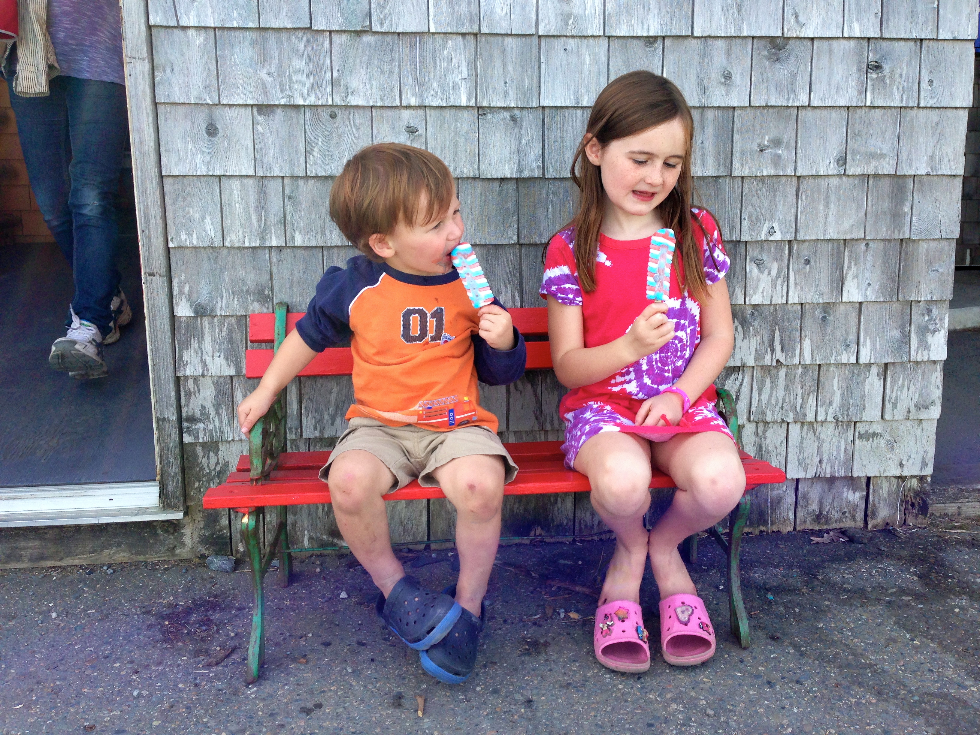 a boy and girl sitting on a bench eating popsicles